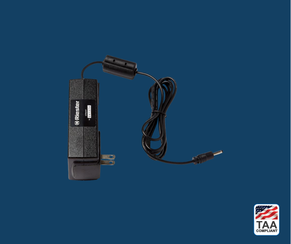 Riester ri-charger L desktop charger, positioned centrally on blue background; TAA Compliant box featuring American flag visible in lower right corner.