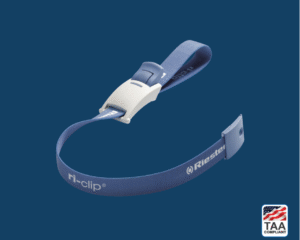 Small blue medical clip positioned in center on blue background; TAA Compliant box with American flag in lower right corner